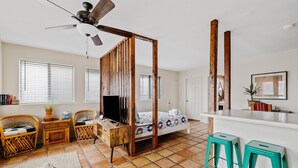 Ein Zimmer mit einem Bett, zwei Korbstühlen, einem kleinen Holztisch, einem Deckenventilator und einem Fenster mit Jalousien.