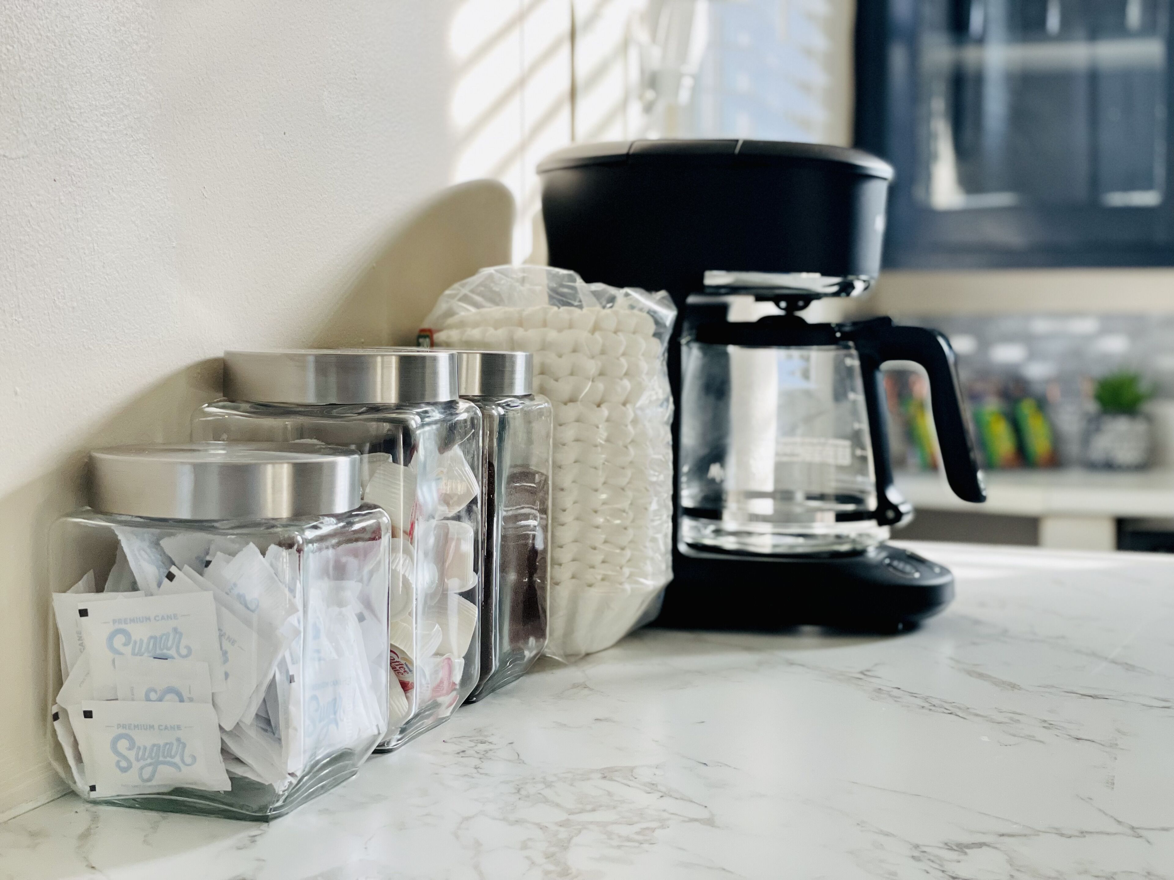 A kitchen counter with a coffee maker, a white cloth, and glass containers with sugar packets.