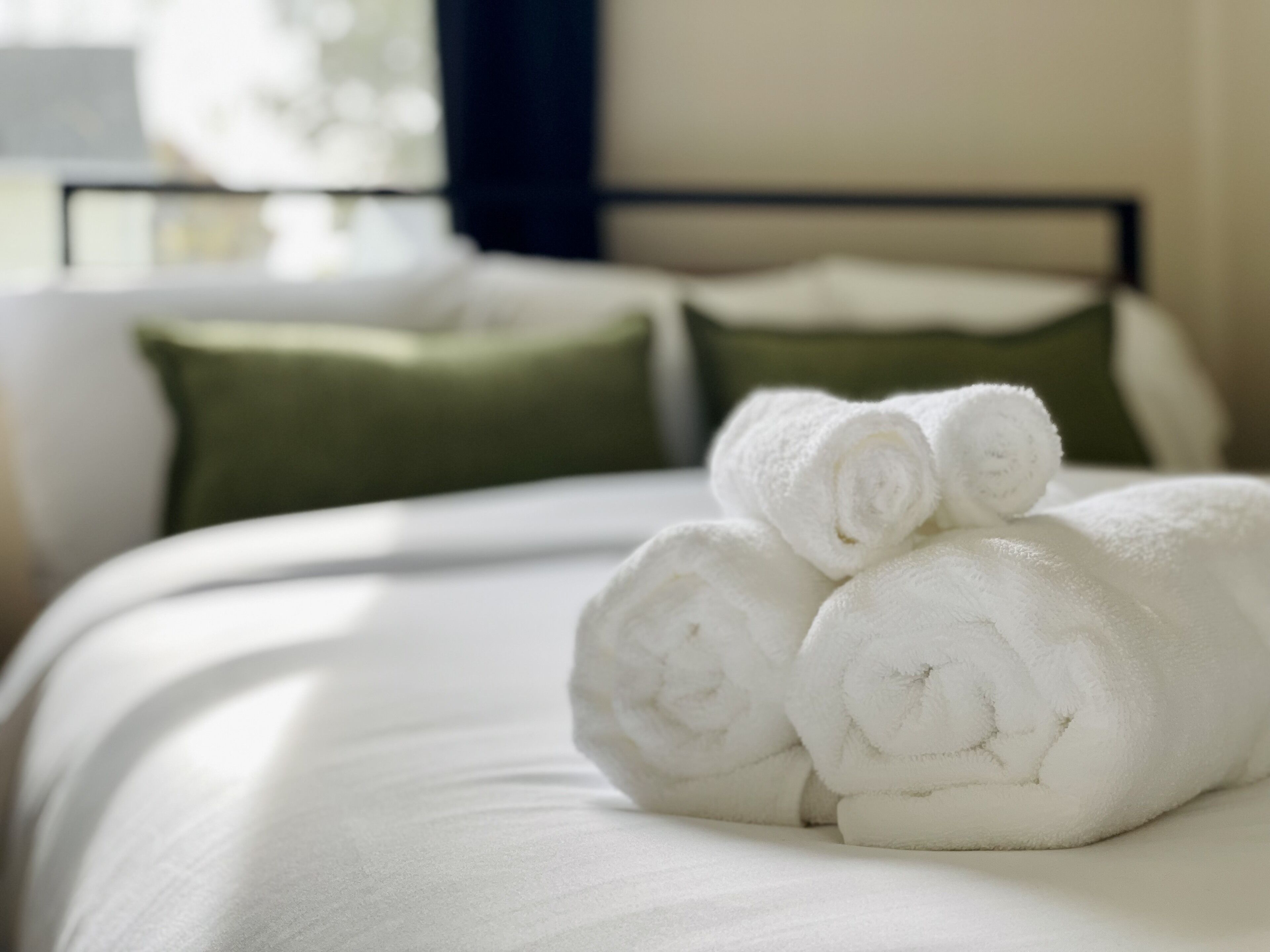 En pænt redt seng med sammenrullede hvide håndklæder og grønne puder i et hotelværelse.