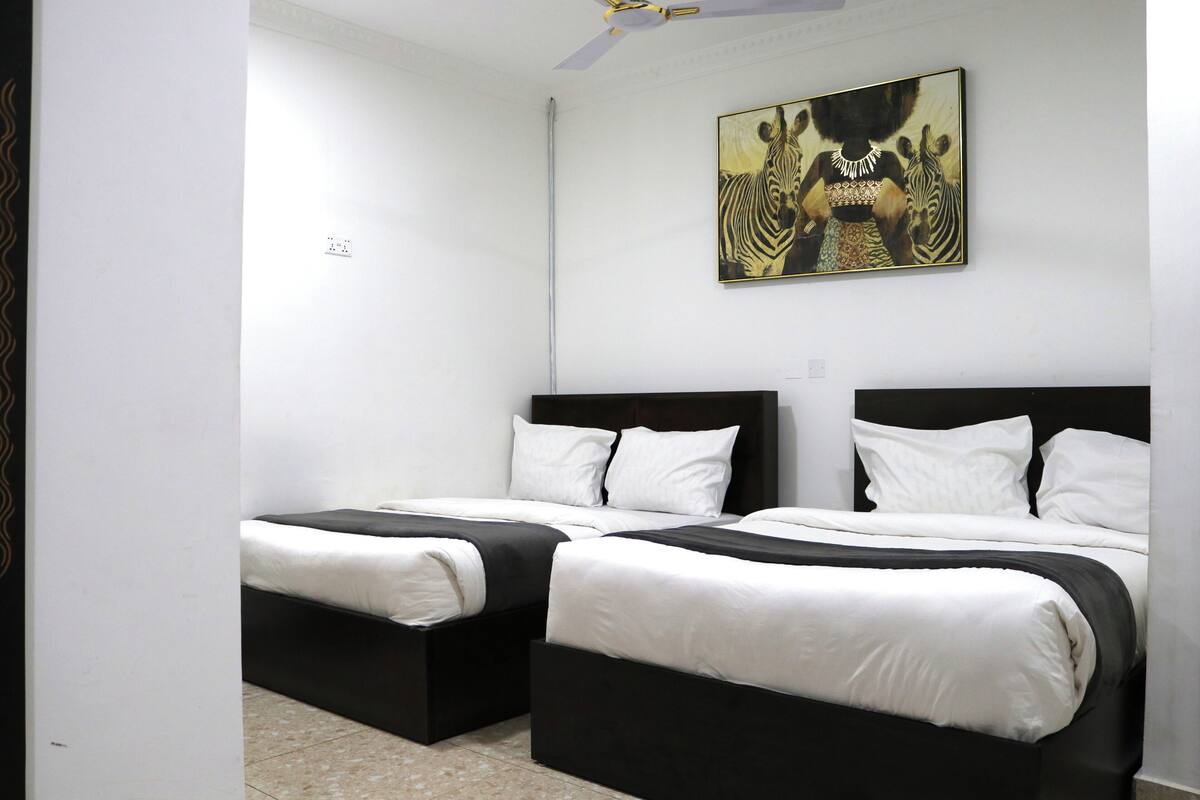 Habitación de hotel con dos camas, ropa de cama blanca, ventilador de techo y un cuadro de cebra en la pared.
