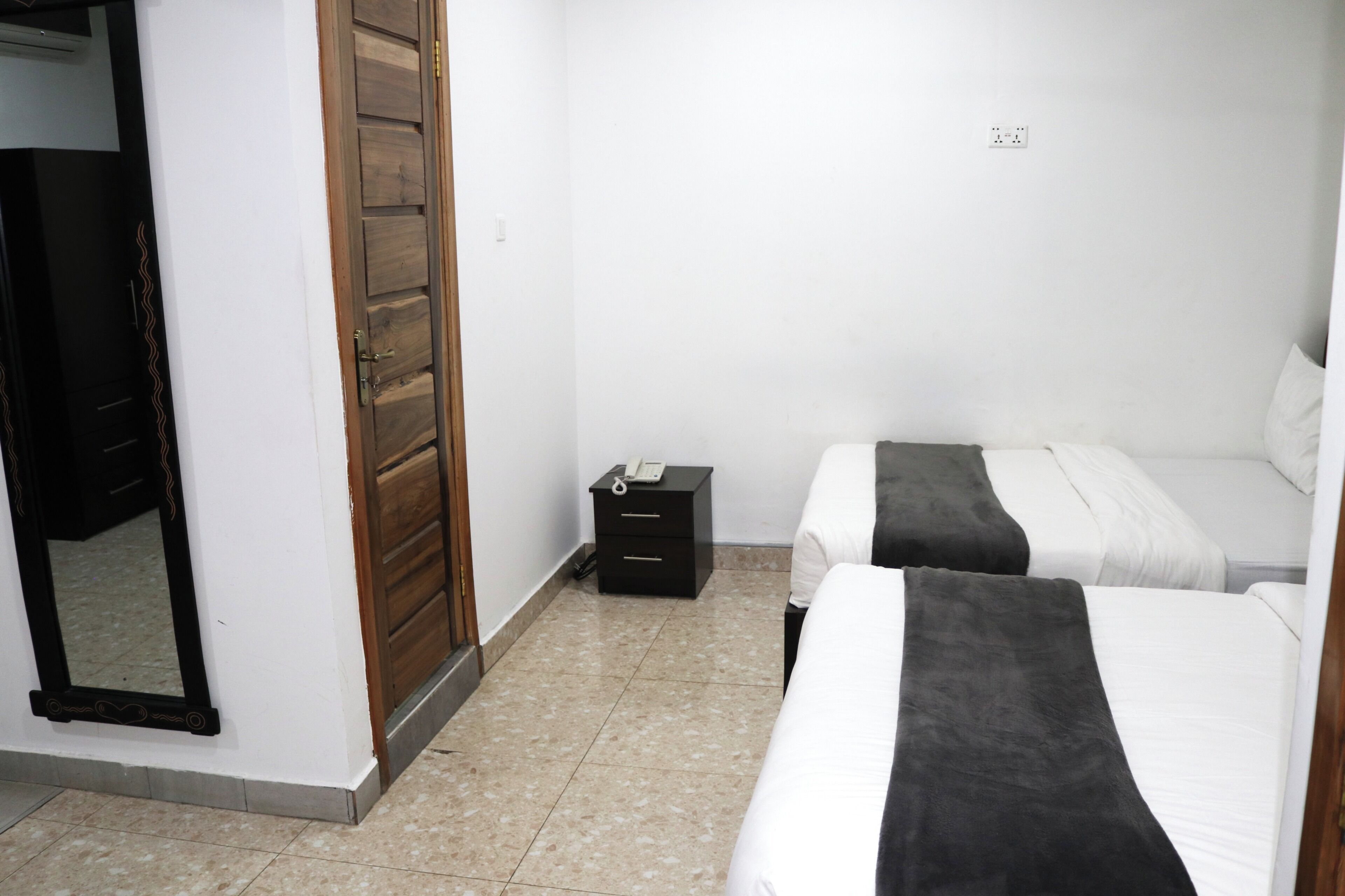 A bedroom with two beds, a wooden wardrobe, a bedside table with a phone, and a mirror.