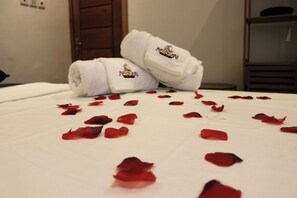 Un lit avec un drap blanc, deux serviettes enroulées ornées d’un logo, et des pétales de rose éparpillés.