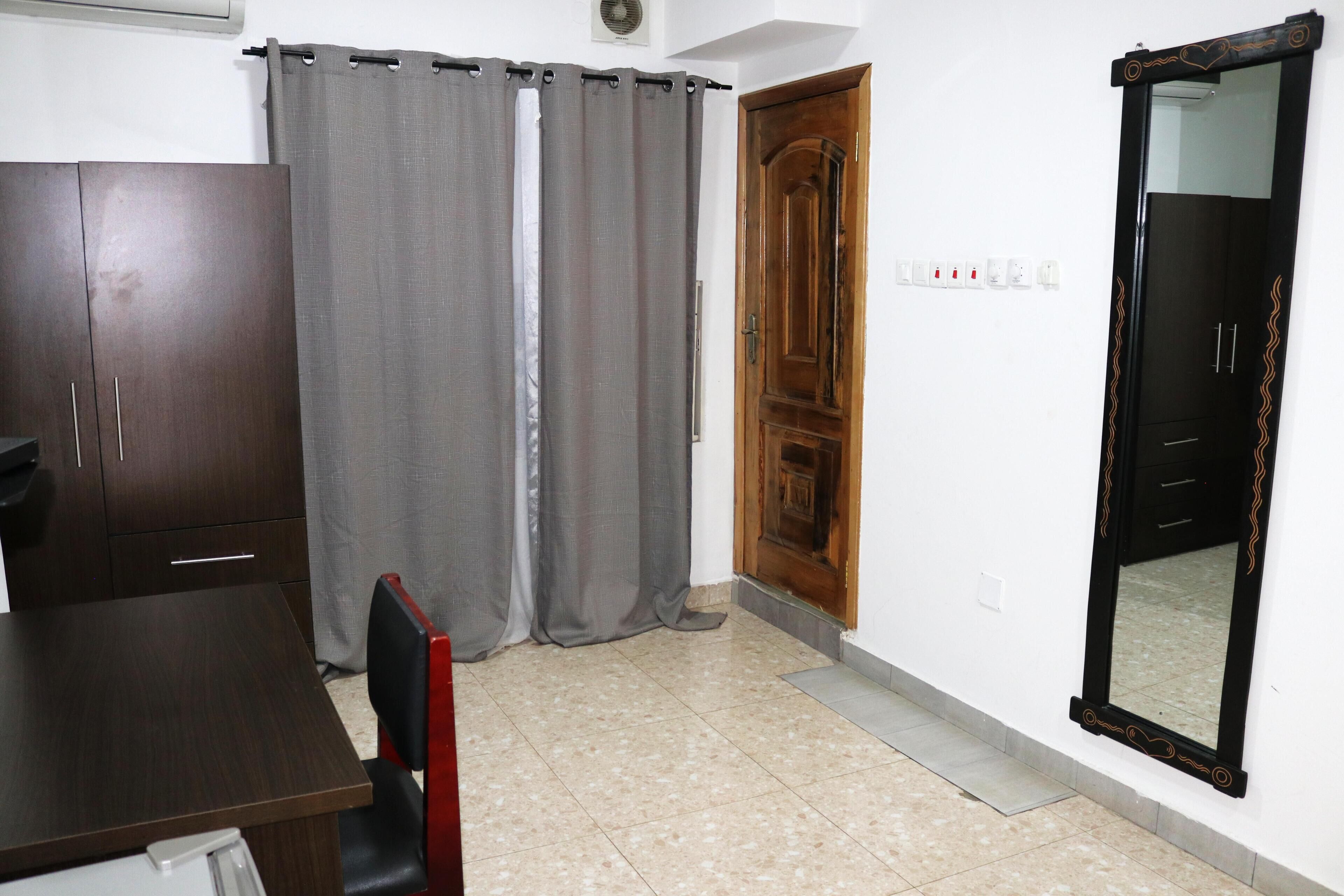 Una habitación con un armario de madera, una mesa con una silla, cortinas grises, una puerta de madera y un espejo fijado a la pared.