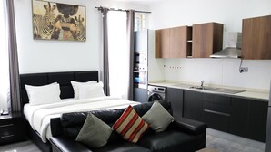 A modern hotel room with a black leather sofa, a bed with white bedding, a kitchen area with wooden cabinets, and a zebra artwork on the wall.