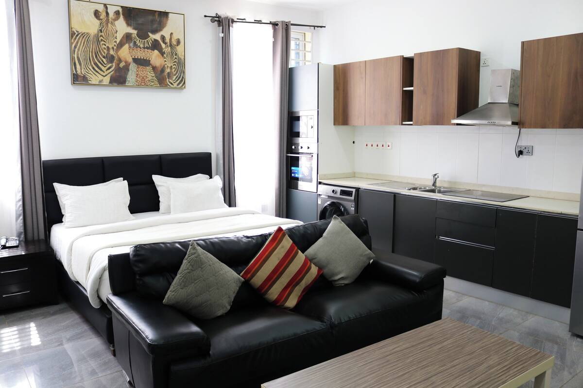 Una habitación de hotel moderna con sofá de cuero negro, cama con ropa de cama blanca, zona de cocina con armarios de madera y una obra de arte con estampados de cebra en la pared.
