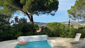 A swimming pool with a white floating bench and lounge chairs, surrounded by lush greenery and a scenic view of hills. - 100m2 house with private pool (Sainte-Maxime)