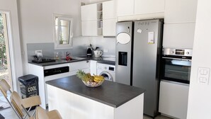 A modern kitchen with white cabinets, a stainless steel refrigerator, a built-in oven, a coffee maker, and a washing machine. There is a fruit bowl on the island and wooden chairs. - 100m2 house with private pool (Sainte-Maxime)