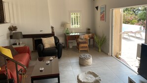 A living room with a sofa, coffee table, and a dining area with a table and chairs. There is a window with a view of greenery outside. - 100m2 house with private pool (Sainte-Maxime)