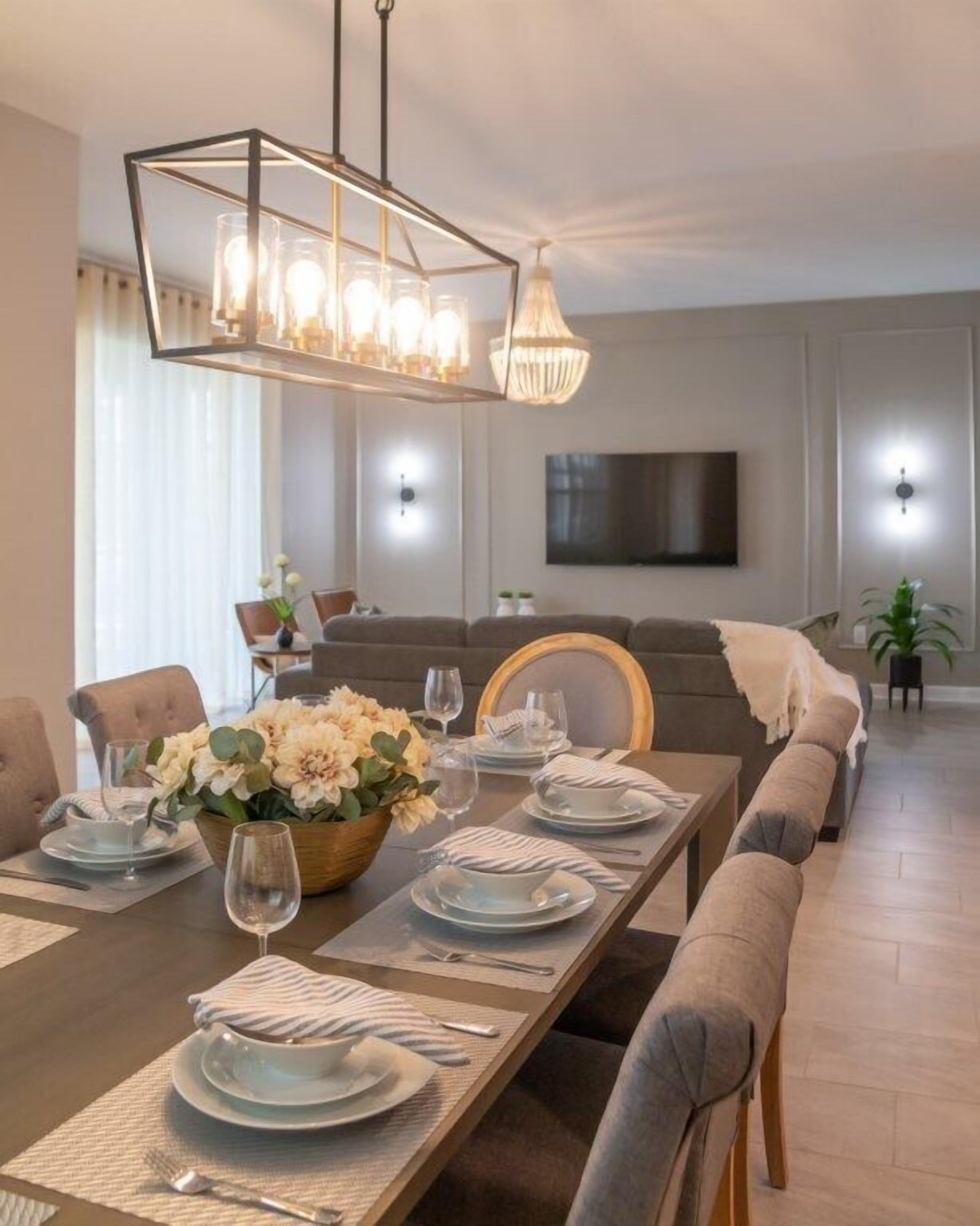En moderne spisestue med et langt bord dækket til seks personer, en lysekrone og en sofa i baggrunden.