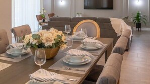A modern dining room with a long table set for six, a chandelier, and a sofa in the background.