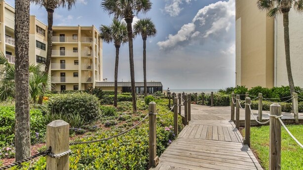 A wooden boardwalk leading to a building with balconies, surrounded by palm trees and a garden.
