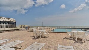 A rooftop pool area with lounge chairs and tables, a pool, and a view of the ocean.