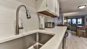 A modern kitchen with a stainless steel sink, white tiled backsplash, and white cabinets. There is a dining area with a table and chairs visible in the background.
