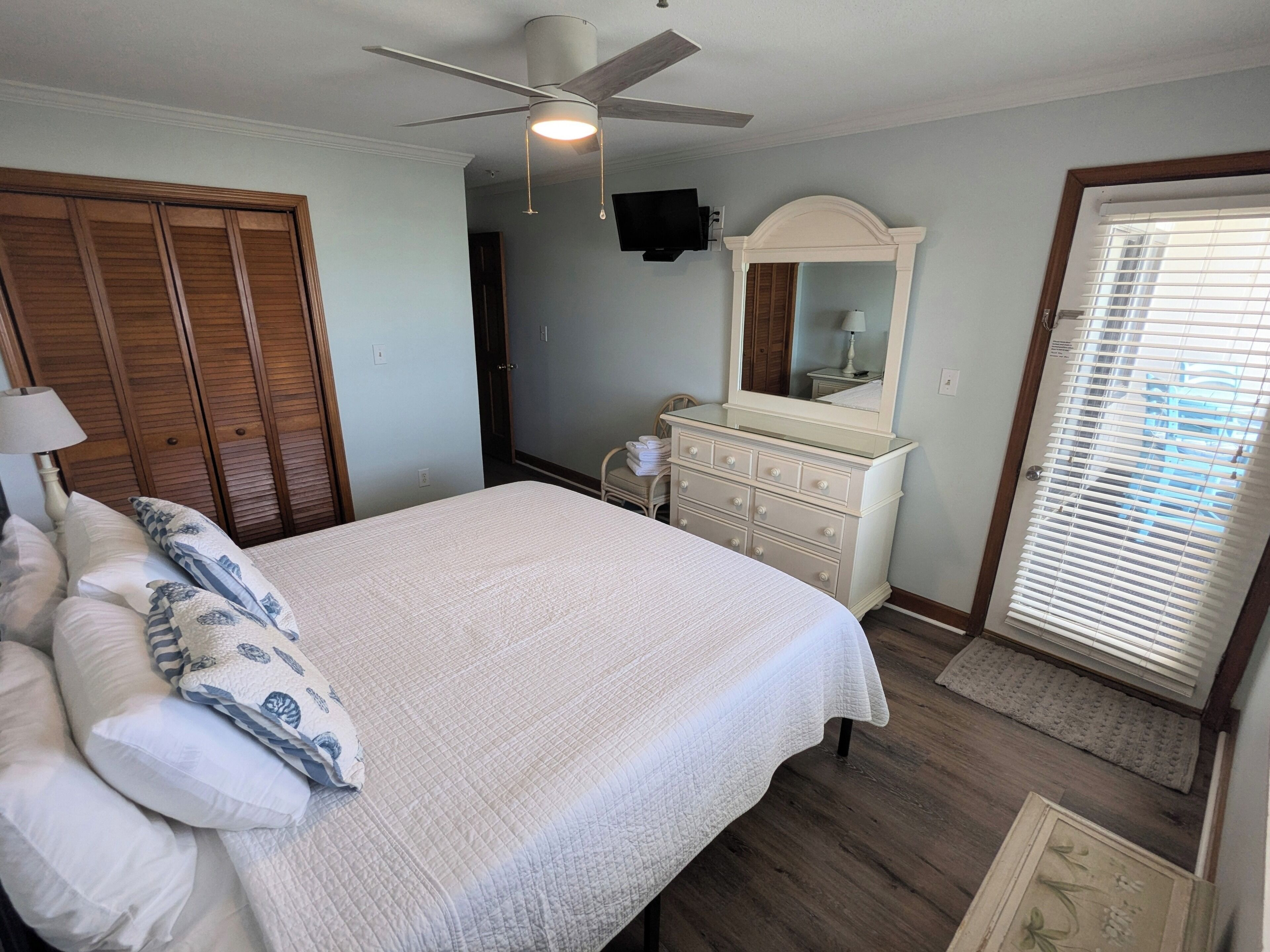 A bedroom with a bed, a dresser, a ceiling fan, a mirror, and a door with blinds.