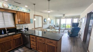 A spacious kitchen with wooden cabinets, a central island, and a dining area with a blue chair. - Spacious oceanfront condo with beachfront pool and elevator! (Surfside Beach)
