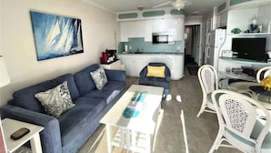 A modern living room with a sofa, coffee table, and chairs. A kitchen with white cabinets and a microwave is visible in the background. - Admiral’s Row 203: Romantic Oceanfront Retreat with Bunk Beds (Hilton Head Island)