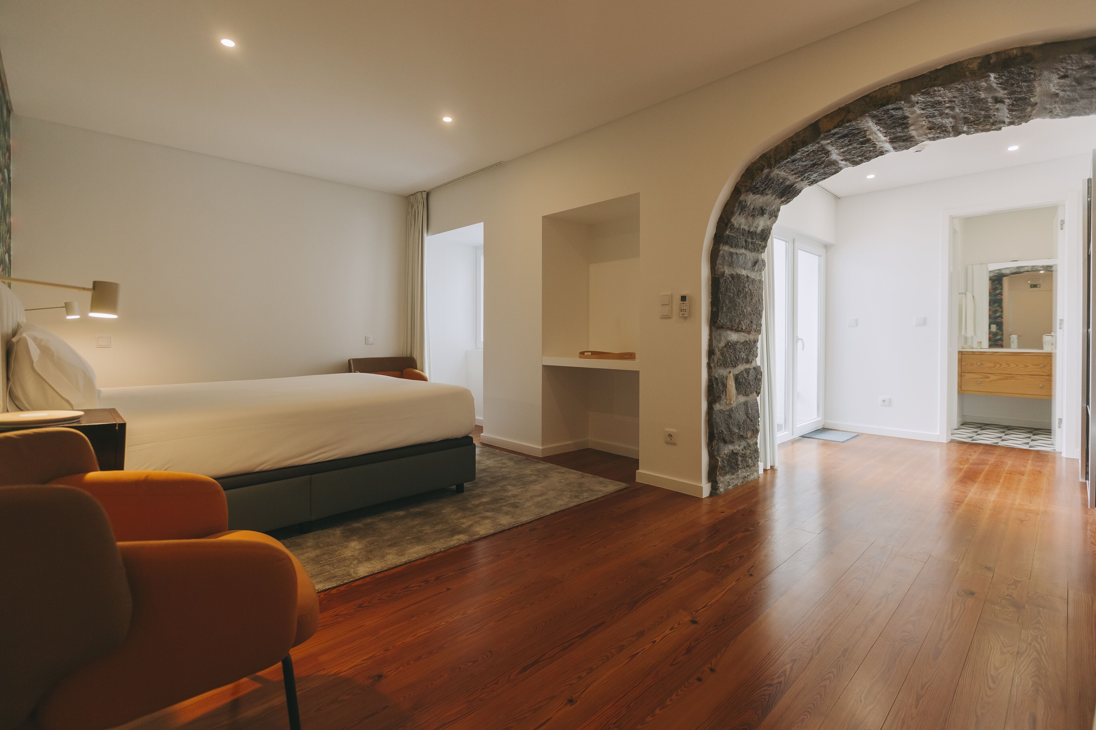 A bedroom with a bed, a desk, and a chair. There is a stone archway leading to a bathroom.