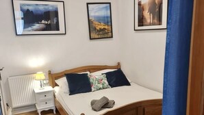 Une chambre avec un lit en bois, une table de chevet blanche et un radiateur. Trois tableaux encadrés sont accrochés au mur.