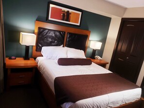 Ein Hotelzimmer mit einem großen Bett, zwei Nachttischen, einem Beistelltisch, einem gerahmten Bild über dem Bett und einer Tür.