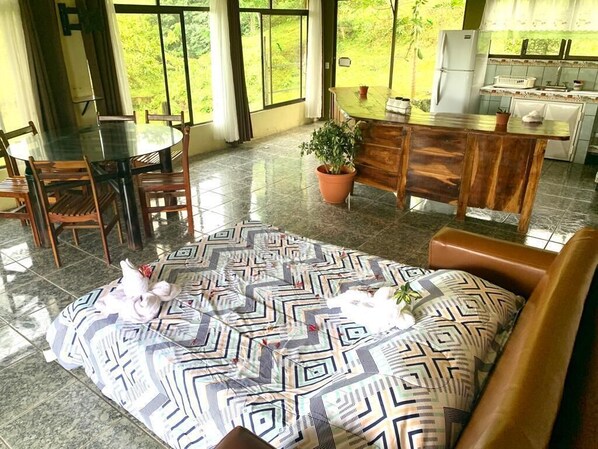 A spacious room with a round glass table and wooden chairs, a patterned sofa, a potted plant, and a kitchen area visible through the windows.