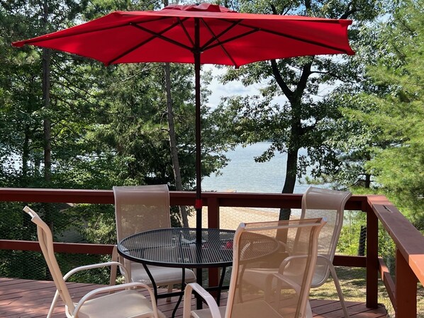 A deck with a round table and four chairs, an umbrella, and a view of a lake surrounded by trees.