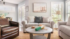A living room with a beige sofa, a brown leather armchair, and a round coffee table. There is a painting of lighthouses on the wall, and a lamp on a side table. - Cheerful Sound Side Condo (Kill Devil Hills)