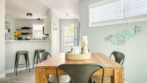 A dining area with a wooden table, four chairs, a decorative wall piece, and a kitchen in the background. - Cheerful Sound Side Condo (Kill Devil Hills)