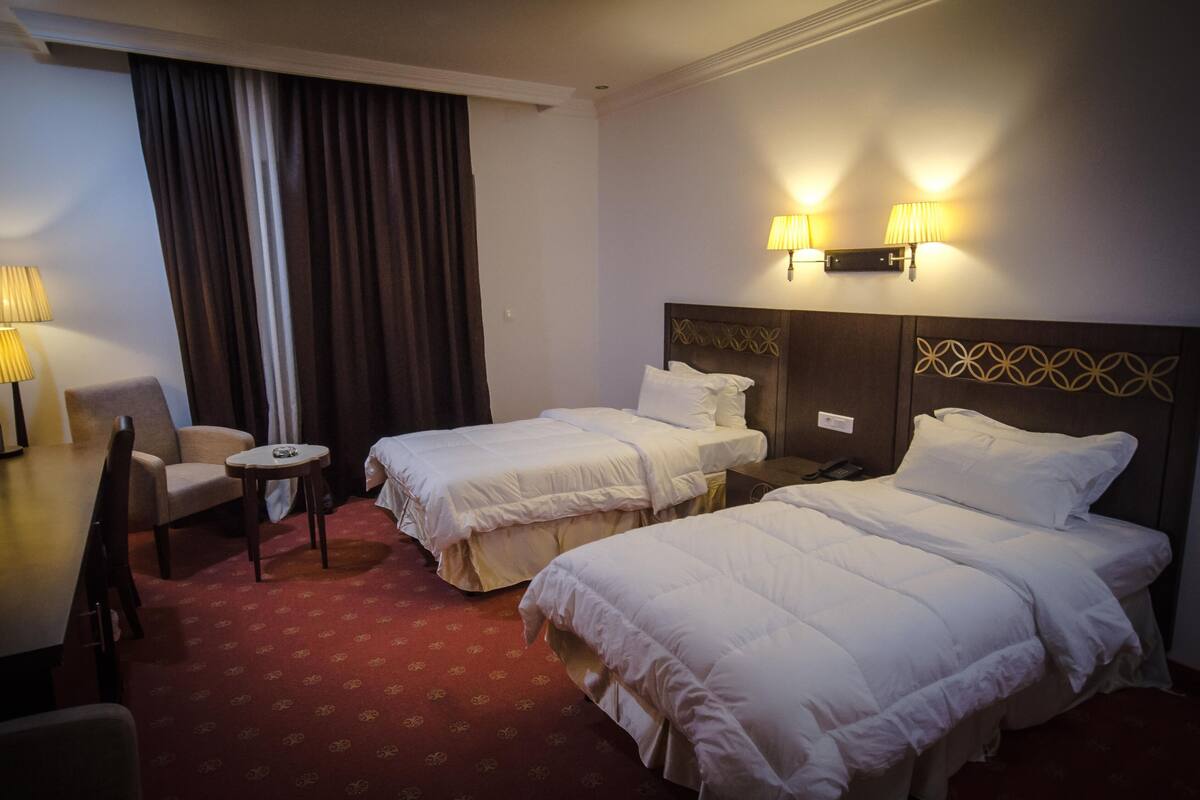 A hotel room with two beds, a small table, a chair, and two wall-mounted lamps.