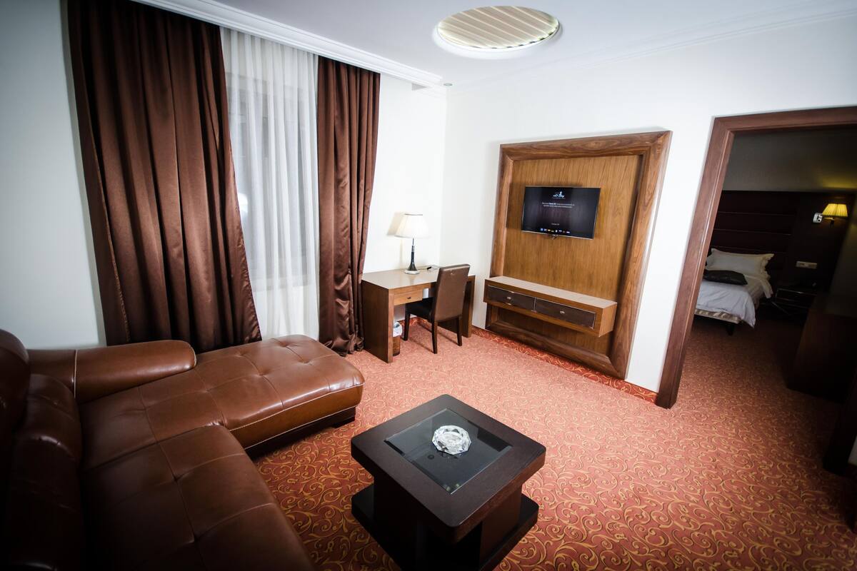 A hotel room with a brown leather sofa, a small table with a glass top, a TV on a wooden cabinet, and a bed with white bedding.