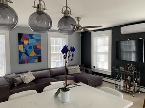 A modern living room with a sectional sofa, a dining table, pendant lights, a ceiling fan, a large TV, and a colorful abstract painting. - Luxury modern house  off Thames Street (Newport)