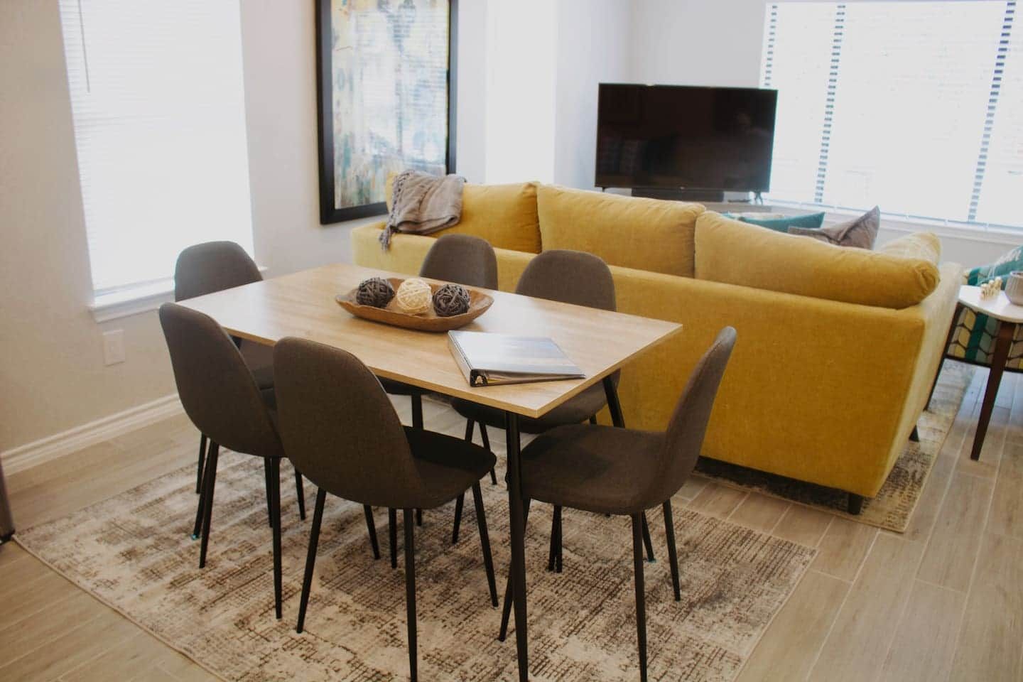 Ein Wohnzimmer mit einem gelben Sofa, einem Holztisch mit Notizbuch und Dekorationskugeln sowie einem Fernseher im Hintergrund.