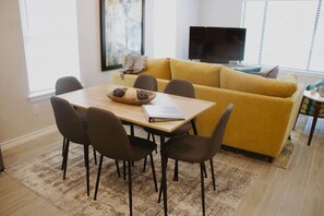 Una sala de estar con un sofá amarillo, una mesa de centro de madera con un cuaderno y bolas decorativas, y un televisor al fondo.