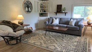 Un salon avec un canapé gris, un tapis à motifs, une table basse en bois, un fauteuil en osier et une étagère murale agrémentée d’objets décoratifs.
