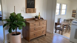 Une commode en bois avec des tiroirs, une grande plante en pot, un tableau noir sur lequel est écrit « Profitez-en ! », et une petite table avec une bouteille et une plante.