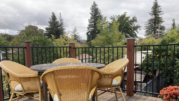Une terrasse en bois, agrémentée de chaises et d’une table en osier, entourée d’une rambarde en métal noir et de verdure.