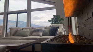 Ein gemütliches Wohnzimmer mit großem Fenster, Kamin und einer Couch mit vielen Kissen.