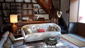 Una sala de estar acogedora con un sofá blanco, cojines estampados, una mesa de centro de vidrio y una estantería de madera llena de objetos varios.