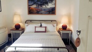 Una cama bien hecha con una colcha estampada, dos lámparas de noche, un cuadro de paisaje enmarcado sobre la cama y una puerta de madera con un gancho para llaves.