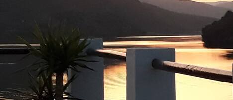 Un balcon agrémenté d’un pot de fleurs, d’une balustrade et offrant une vue sur un lac paisible, avec des montagnes en arrière-plan.