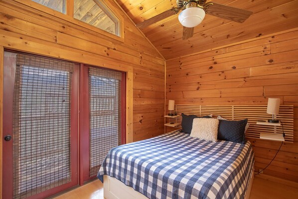 A wooden cabin bedroom with a bed, two bedside tables, a ceiling fan, and a window with blinds.