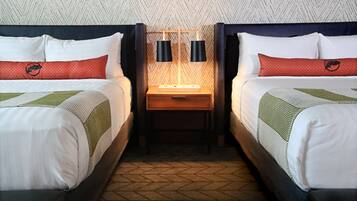 Quarto de hotel com duas camas, cada uma com cabeceira, mesa de cabeceira e luminária, além de um tapete estampado.