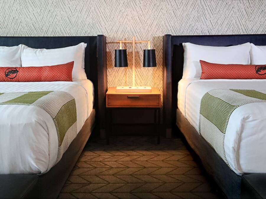 Quarto de hotel com duas camas, cada uma com cabeceira, mesa de cabeceira e luminária, além de um tapete estampado.