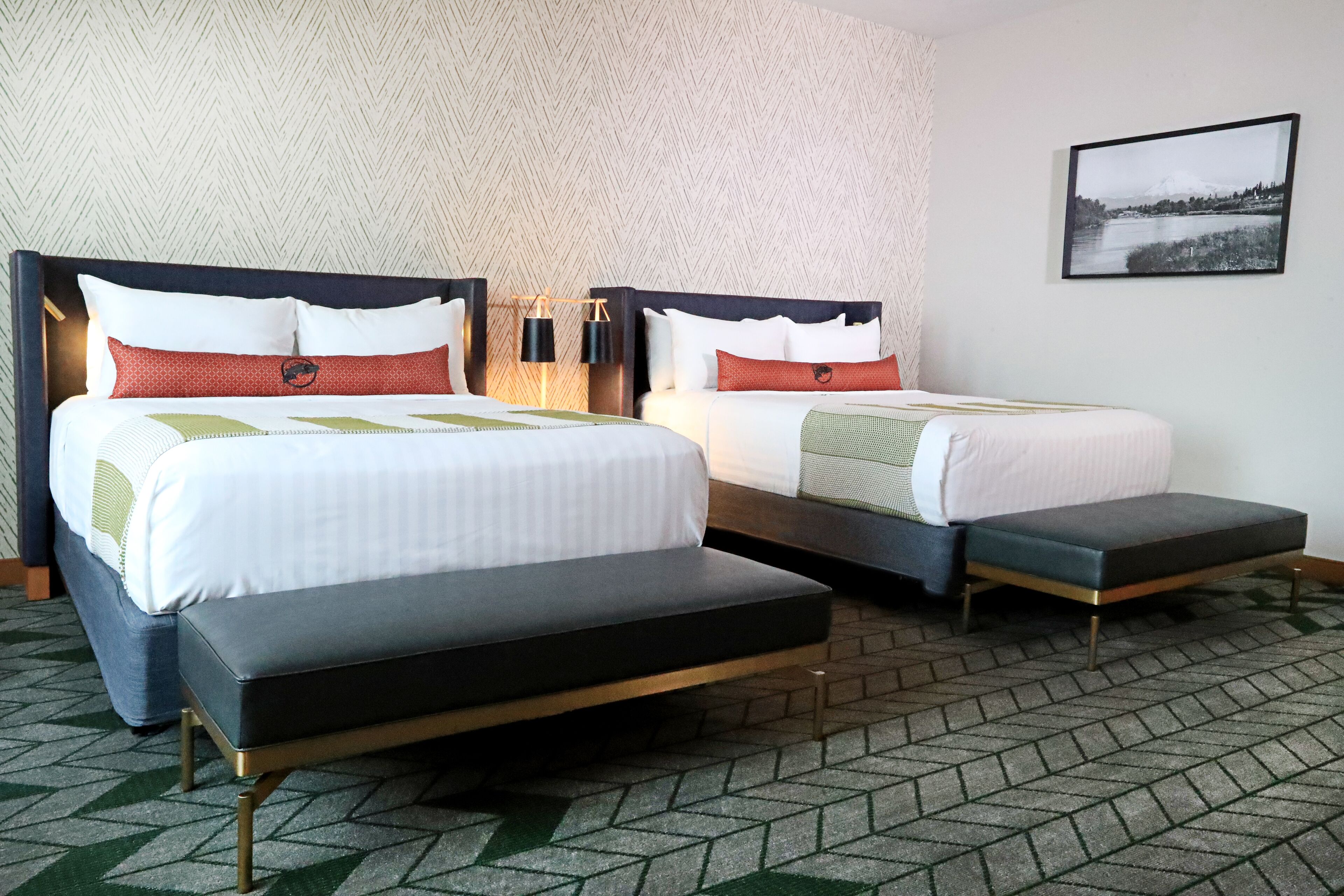 A hotel room with two beds, a bench, a patterned carpet, and a framed picture on the wall.