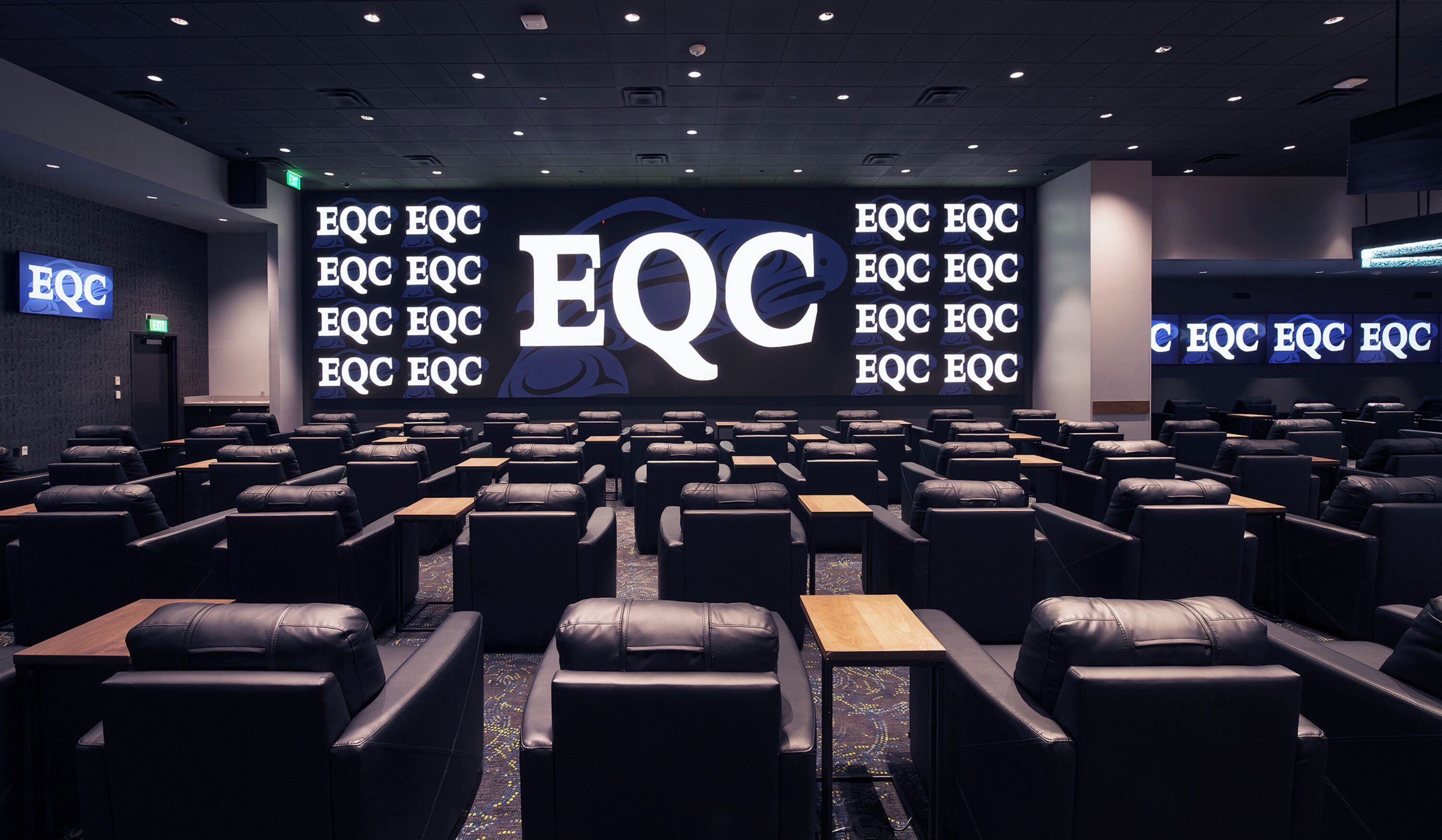 Een congrescentrum met rijen zwarte lederen stoelen en tafels, een groot scherm waarop herhaaldelijk 'EQC' wordt weergegeven, en een 'FOC'-bord aan de linkerkant.