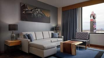 A modern hotel room with a grey sofa, two side tables with lamps, a wooden coffee table, a patterned armchair, and a large window with a view of a cityscape and a bridge.