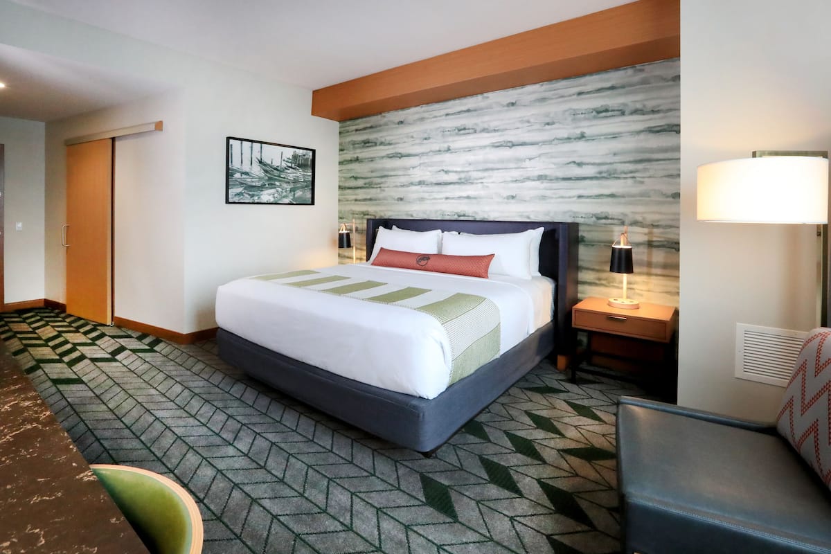 Ein Hotelzimmer mit einem großen Bett, einem Nachttisch, einer Lampe, einem Sessel und einem gemusterten Teppich.