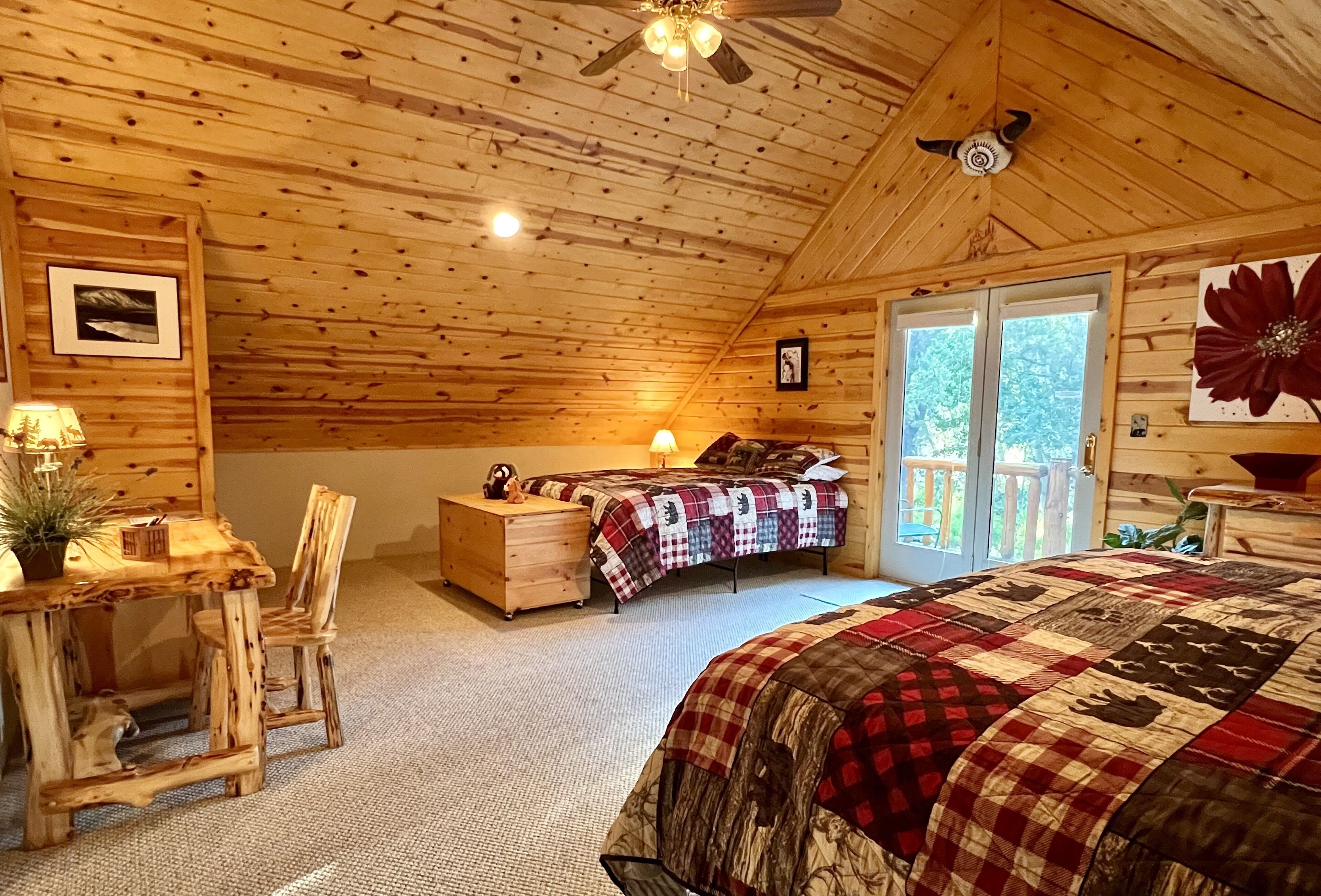 Un dormitorio de cabaña acogedor, con techo de madera, cama con colcha a cuadros, escritorio con lámpara y vista al exterior a través de una puerta corrediza.