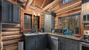 Une cuisine moderne dans un chalet en bois, avec des meubles sombres, un îlot central et une grande fenêtre offrant une vue sur l’extérieur.