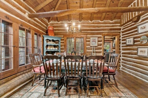 A log cabin interior with a dining table surrounded by wooden chairs, a chandelier, and a large window.