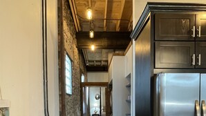 A narrow hallway with wooden floors and a stone wall on one side, leading to a kitchen area with a stainless steel refrigerator and wooden cabinets. - Historic 1 bed 1 bath in 1920's Chevy (Mountain Home, AR, 72653)